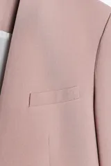 Blazer color rosa palo, sin solapa, de corte recto y manga larga. Presenta bolsillos de ojal en el delantero y cierre frontal con gancho metálico.