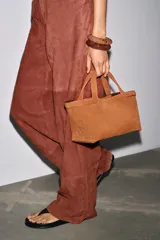 Bolso mini shopper color marrón de serraje, con asa de mano y asa bandolera extraíble. Tiene detalle de pespunte en el corte, bolsillo interior y cierre mediante mosquetón.