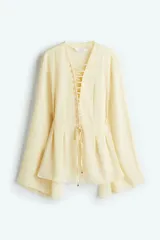 Blusa color crema de gasa con escote en V y cordones cruzados. Tiene mangas largas acampanadas y pliegues en la cintura.
