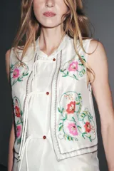 Top blanco de cuello mao y sin mangas, con bordados florales en el frente y cierre con botones.