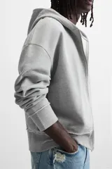 Sudadera gris con capucha, corte holgado, cierre frontal con cremallera, bolsillos laterales ocultos y terminaciones irregulares.