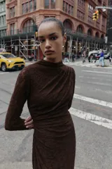 Vestido midi de manga larga, color marrón oscuro, confeccionado en tejido de encaje o punto con patrón floral sutil. Presenta cuello perkins y drapeado asimétrico en el torso con frunces laterales.