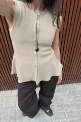 Top de punto acanalado en color beige, sin mangas y con cuello redondo. Presenta un cierre frontal con botones pequeños y un bajo con acabado en peplum que aporta volumen.