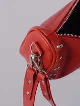 Bandolera color rojo de cuero con tachas plateadas. Tiene dos asas desmontables, una corta con tachas para llevarla en la mano o al hombro y una larga para llevarla al hombro o cruzada. Tiene bolsillo interior con cierre y forro.