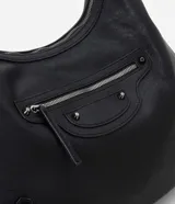 Cartera tipo hobo grande de material sintético negro con textura que imita cuero. Presenta un asa de hombro fija con hebillas metálicas, cierre principal superior de cremallera y un bolsillo frontal con cremallera y detalles de tachuelas decorativas.