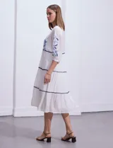 Vestido midi blanco con bordados florales azules en el escote, mangas y cintura. Tiene cuello en V, mangas 3/4 con botón y falda escalonada con detalles de ribetes azules.