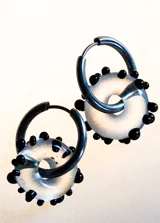 Par de aros colgantes con forma de dona transparente con pequeñas bolitas negras adheridas, y aro de acero quirúrgico.