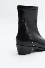 Bota corta de cuero sintético color marrón oscuro, con caña elástica y puntera afilada. Presenta una hilera de tachas metálicas doradas en el lateral exterior y un taco bajo y estable.