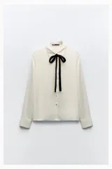 Camisa color crema de satén con cuello solapa, manga larga y lazo negro contrastante. Cierre frontal con botones.