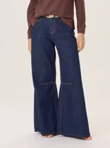 Pantalón de jean azul oscuro, tiro alto, con corte ancho y acampanado (flare) desde la rodilla. Presenta costuras decorativas a contratono (naranja/marrón) y un detalle de pinzas o costura horizontal marcada debajo de cada rodilla.