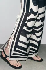 Pantalón de tiro medio con cintura elástica y pierna ancha. Presenta un estampado de líneas onduladas en blanco y negro, y aberturas laterales ajustables con lazadas.