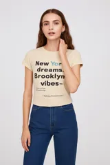 Remera color crema de algodón con cuello redondo y mangas cortas. Estampa frontal con texto "New York dreams. Brooklyn vibes" en color negro y verde.