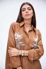 Camisa marrón de manga larga con puños anchos, cuello camisero y botones frontales. Presenta bordados florales en blanco con lentejuelas y mostacillas en la parte delantera.