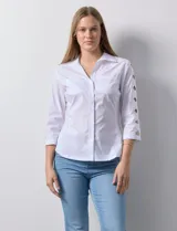 Camisa blanca de algodón con cuello clásico, solapa y escote en V. Tiene mangas tres cuartos con detalle de aberturas cruzadas. Presenta dos bolsillos delanteros aplicados y laterales en tejido modal.