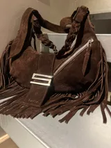 Bolso tipo hobo de cuero gamuza color marrón oscuro, con flecos largos que rodean la base. Presenta un asa corta trenzada y un cierre diagonal con cremallera metálica. Incluye un detalle de correa frontal con hebilla metálica plateada y la inscripción "SOFIA" grabada.