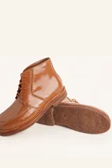 Bota de cuero vacuno con acabado charol, color marrón oscuro. Presenta un diseño de caña corta con cordones frontales y costuras decorativas en la puntera. La suela es de goma tipo crepe.