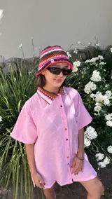 Camisa de lino color rosa claro, de manga corta y corte holgado. Posee cuello estilo polo con tejido a crochet en tonos marrón, rosa y blanco, y botones frontales multicolor.