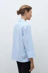 Camisa de popelina celeste con finas rayas blancas verticales, cuello camisero y abertura frontal con botones joya. Manga larga con puño ancho.