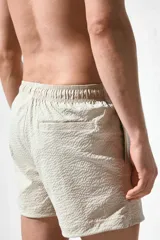 Short de baño color beige con textura de sirsaca, cintura elástica con cordón ajustable, bolsillos laterales y trasero ribeteado, aberturas laterales cortas y suspensor interior de malla.