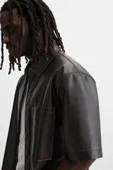 Sobrecamisa negra de manga corta confeccionada en tejido efecto piel, con cuello solapa, bolsillo frontal de parche y cierre frontal con botonadura oculta.