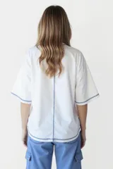 Remera blanca de corte holgado con cuello redondo y mangas cortas. Presenta un pequeño estampado de una flor amarilla en el centro del pecho y detalles de costura en azul en cuello, mangas y ruedo.