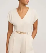 Enterito corto de mujer color beige claro, con escote en V, mangas cortas tipo capa, trabillas en la cintura y pliegues frontales. Incluye un cinturón fino trenzado de cuerda.