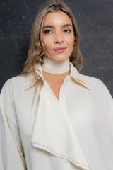 Blusa color crema de poliéster con cuello doble estructurado, volado delantero, lazo regulable en cintura y abotonada al frente.