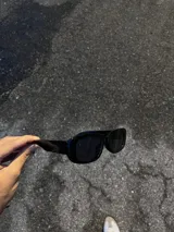 Gafas de sol negras con montura rectangular gruesa y lentes oscuras.