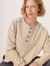 Buzo de manga larga con diseño de bloques de color y textura. El cuerpo principal es marrón, la espalda superior y hombros tienen un panel de tejido a rayas grises y azules, y los hombros superiores son de color beige rosado. Presenta costuras decorativas en rojo que separan los bloques de color.