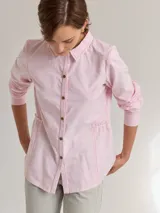 Camisa rosa claro de algodón con cuello clásico, mangas largas y cierre frontal con botones. Presenta un volante en la parte inferior delantera y costuras a contratono en color rojo.