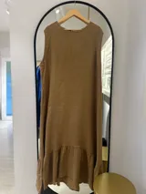 Vestido largo sin mangas color tostado, de tela ligera con textura, corte recto y holgado, con un volante fruncido en el ruedo inferior. El cuello es redondo con borde deshilachado.