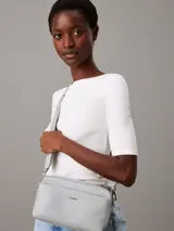 Cartera tipo camera bag de color blanco/gris claro, confeccionada en piel sintética con textura de monograma en el bolsillo frontal. Cuenta con dos correas desmontables: una ancha y otra fina, ambas ajustables. El bolsillo frontal tiene el logo de Calvin Klein en metal plateado.