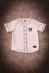 Camisa de béisbol color crema con ribetes negros, cierre frontal con botones negros y bordado de una letra "H" en el pecho.