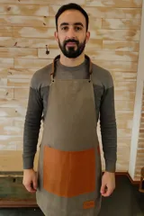 Delantal unisex de lona color gris con bolsillo de cuero marrón y tiras de cuero ajustables.