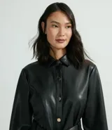 Chaqueta larga de cuero sintético negro, con cuello camisero, cierre frontal con botones, cinturón con hebilla, bolsillos con solapa y puños abotonados.