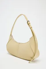 Bolso de hombro color crema, con forma alargada y asa ajustable con hebilla. Tiene pespuntes en el cuerpo, bolsillo interior con cremallera y cierre principal con cremallera y tirador.