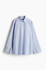 Camisa celeste de algodón, corte amplio, con cuello clásico, abotonadura frontal y mangas largas con hombros caídos y puños con abertura.