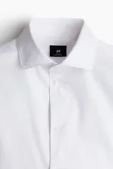 Camisa blanca de algodón con cuello italiano, tapeta lisa, canesú en la espalda y bajo redondeado. Mangas largas con tapeta de manga y botón, y cierre ajustable de botón en los puños. Corte ajustado que realza el contorno del cuerpo creando una silueta entallada.