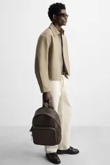 Mochila marrón oscuro de cuero, con compartimento principal con cierre de cremallera, bolsillo interno plano y bolsillo interno con cremallera. Bolsillo frontal con cremallera, parte posterior acolchada con banda de sujeción para equipaje, asa de mano y dos asas de hombro ajustables.