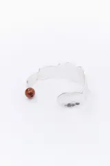 Pulsera rígida plateada con forma geométrica y una cuenta marrón en uno de los extremos.