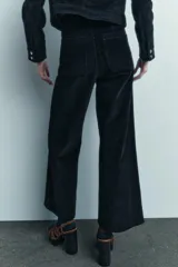 Pantalón ancho de pana color negro, de tiro medio, con trabillas en la cintura, bolsillos delanteros y cierre frontal con cremallera, botón y gancho metálico.