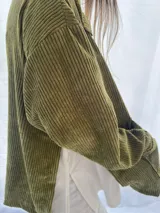 Buzo de pana fina color verde oliva, con diseño de corte holgado y mangas largas. Presenta una textura de cordón vertical característica de la pana y un pequeño parche decorativo en la parte superior de la espalda.