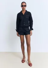 Chaqueta corta de denim azul oscuro, con cuello de solapa, dos bolsillos de parche en el pecho y cierre frontal de botones. Presenta costuras contrastantes y puños abotonados.