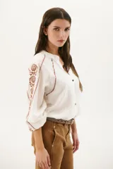 Camisa blanca de manga larga con bordados marrones en hombros y mangas, cuello en V y cierre frontal con botones.