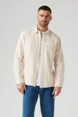 Camisa de manga larga color crema, con cuello clásico, cierre frontal con botones y bolsillo de parche en el pecho izquierdo con logo de la marca.
