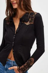 Camisa negra de manga larga con cuello clásico y cierre frontal con botones. Presenta detalles de encaje floral en los hombros y puños.
