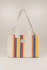 Bolso tote pequeño de algodón a rayas verticales en crudo, amarillo, azul y rojo. Tiene doble asa corta y correa larga regulable para llevar al hombro. Incluye bolsillo para teléfono y bolsillo para llaves.