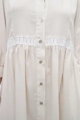 Vestido camisero midi color beige a rayas verticales, con cuello mao, mangas 3/4 abullonadas y festones de algodón en el cuerpo. Cierre frontal con botones.