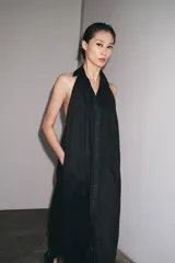 Vestido midi negro de lino, con cuello halter, hombros descubiertos, espalda descubierta, cierre frontal con botones y bajo asimétrico.