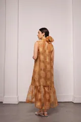 Vestido largo color ocre con estampado de cuadrados dorados, cuello alto con lazo en la nuca y falda con volados. Incluye viso liso al tono.